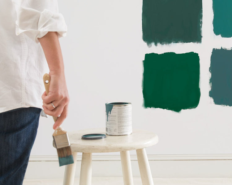 Person in white shirt and blue jeans holding paint brush in front of table with green paint bucket and four square green blocks on the wall